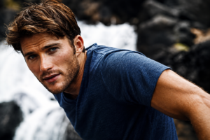 Scott Eastwood in a cinematic outdoor portrait wearing a navy blue shirt, with a dramatic waterfall and rocky landscape in the background.