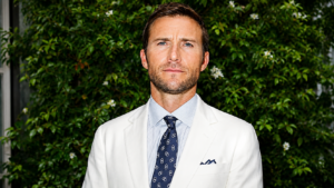 Scott Eastwood in a polished portrait wearing a white suit and patterned navy tie against a lush green outdoor backdrop.