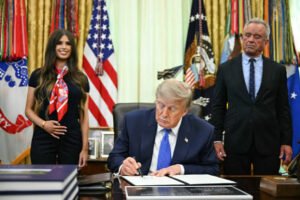 Donald Trump signing an executive document at a desk in the Oval Office, with U.S. flags and officials standing nearby, related to a childhood vaccine policy order.