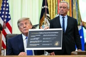 Donald Trump holding a sign titled “Gold Standard Childhood Vaccine Recommendations” in the Oval Office, with U.S. flags and officials in the background.